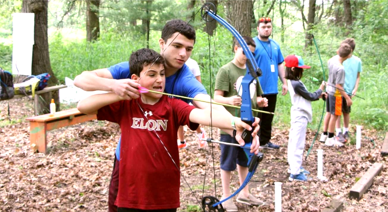 counselor helping a camper shoot a bow and arrow
